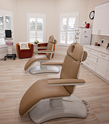 Modern dental clinic with two beige reclining treatment chairs, white cabinetry, windows and a lounge chair.