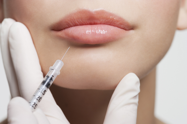 Close-up of a woman receiving a lip filler injection, syringe near lower lip held by gloved hands.