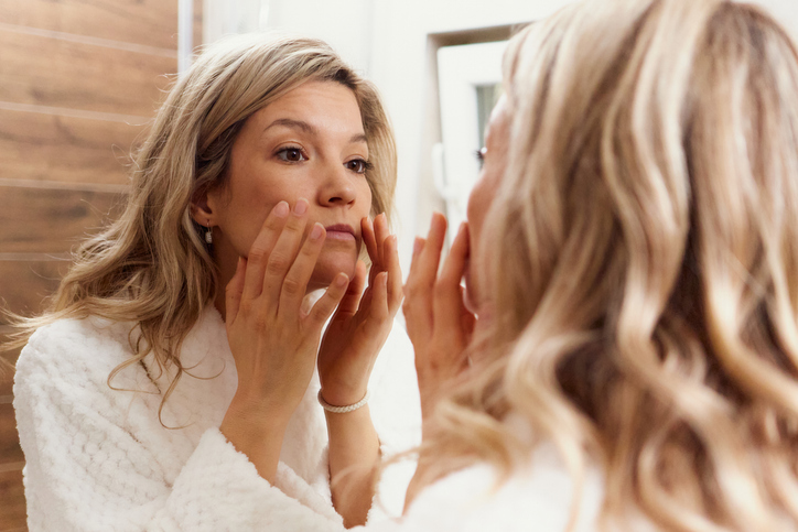 A woman in a white robe looking at her reflection and massaging skincare onto her cheeks.