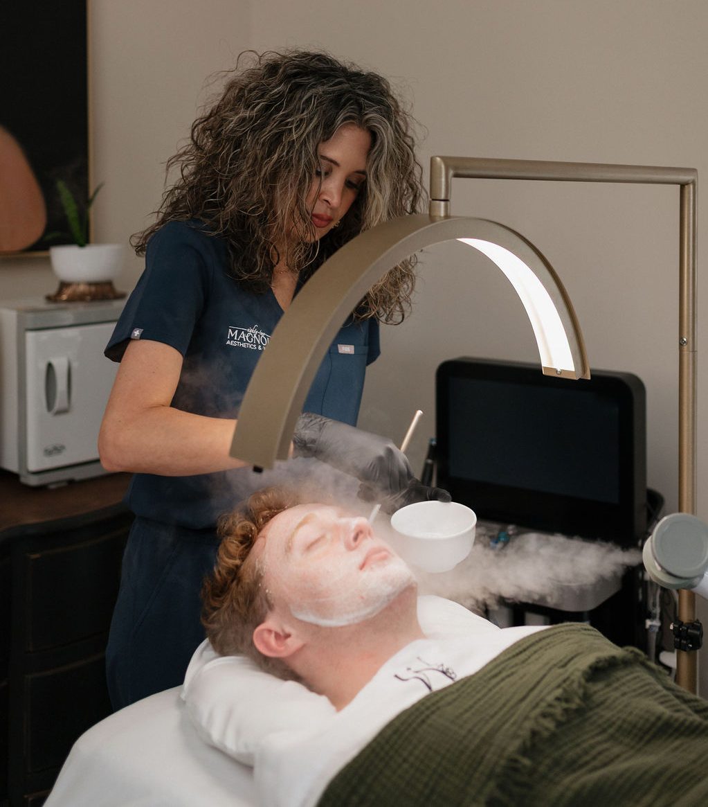 Aesthetician steaming and applying a facial mask to a relaxed man lying under a ring light in a spa treatment room.