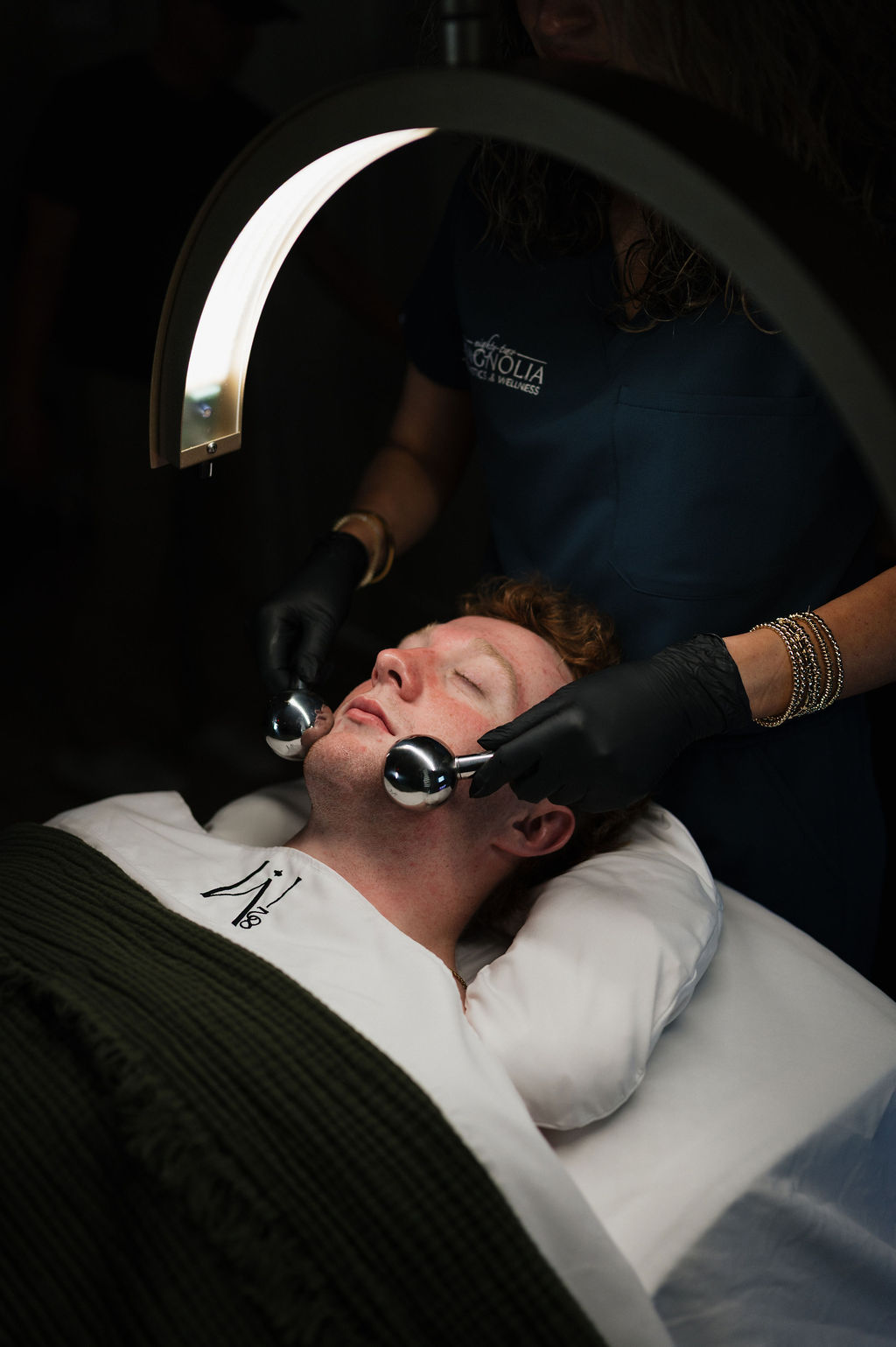 A man receiving a facial massage with metal globe rollers under a curved LED lamp