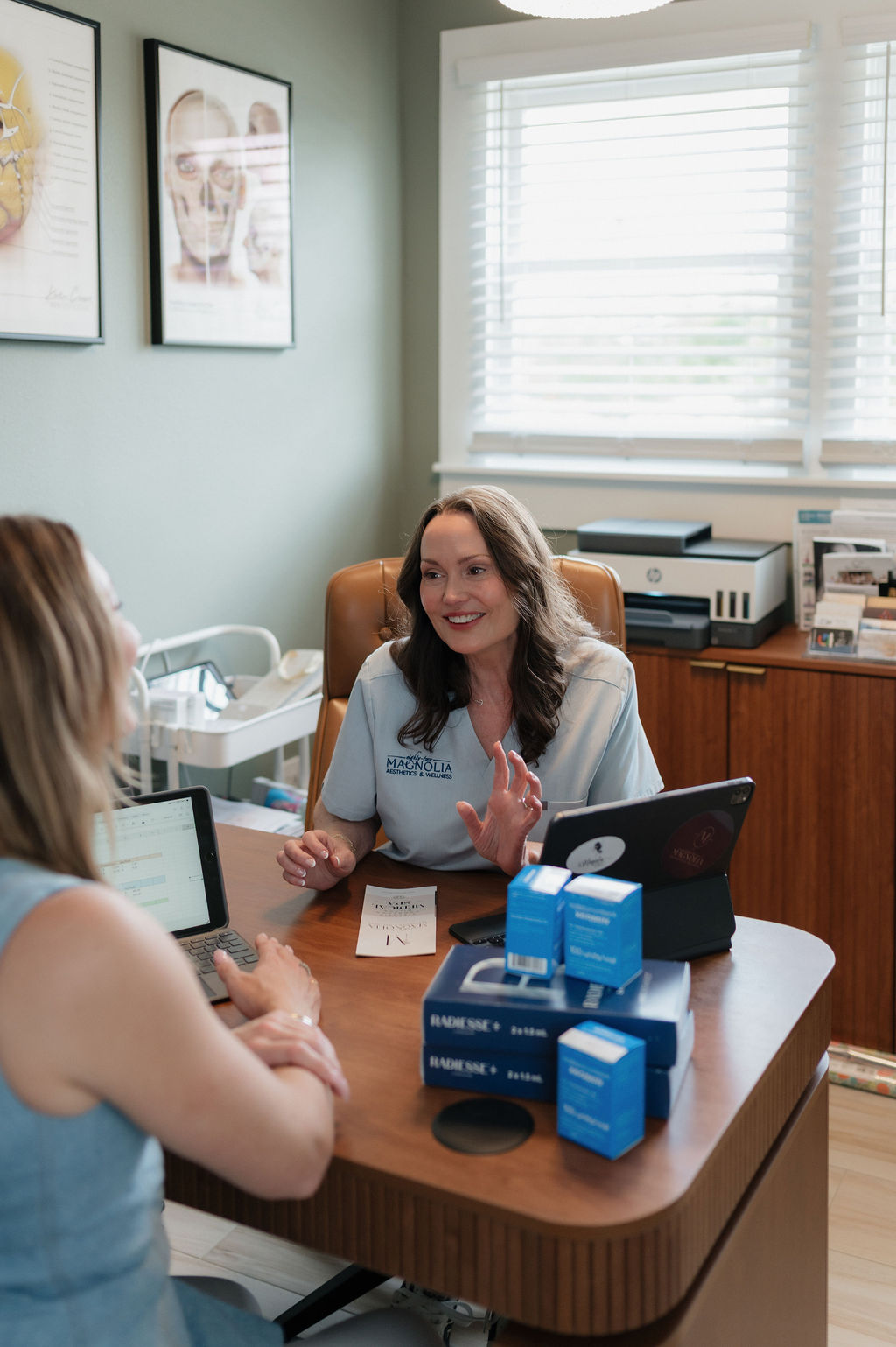 Woman esthetician consulting a client at a medspa desk with skincare product boxes, tablet, and paperwork
