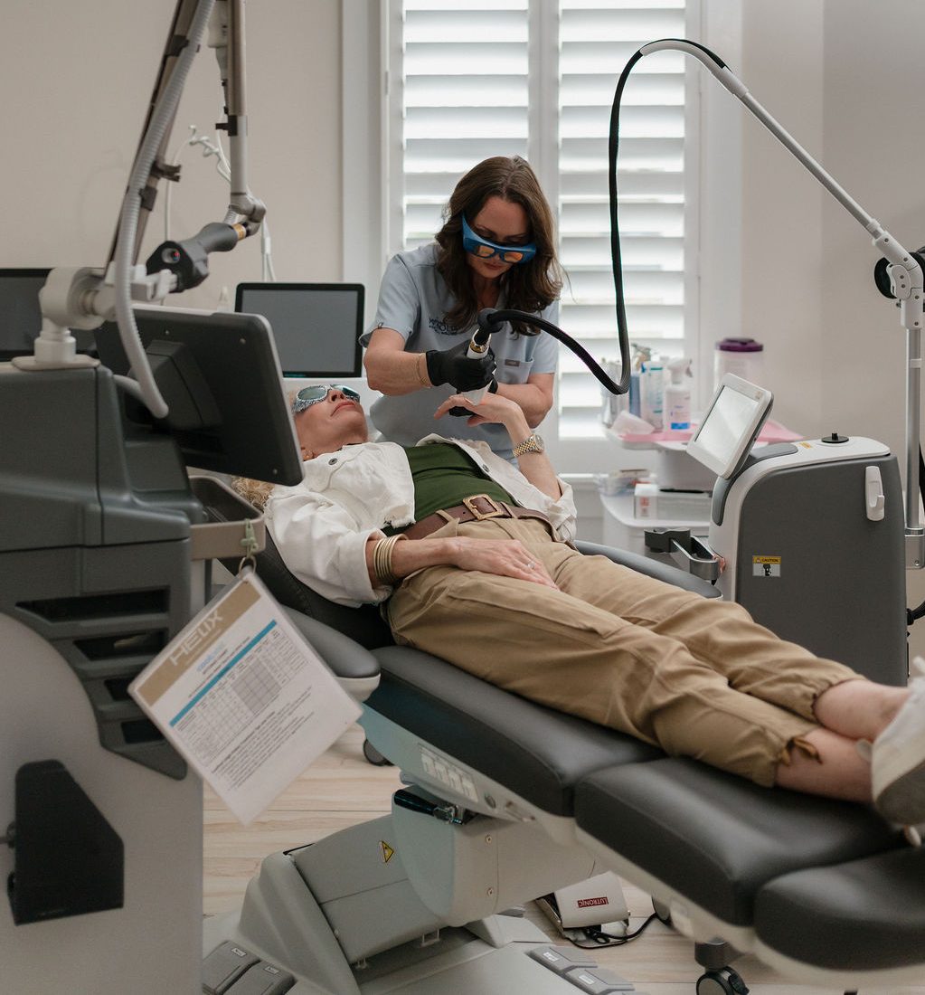 Clinician wearing protective eyewear performing laser skin treatment on a woman reclining in a dermatology clinic