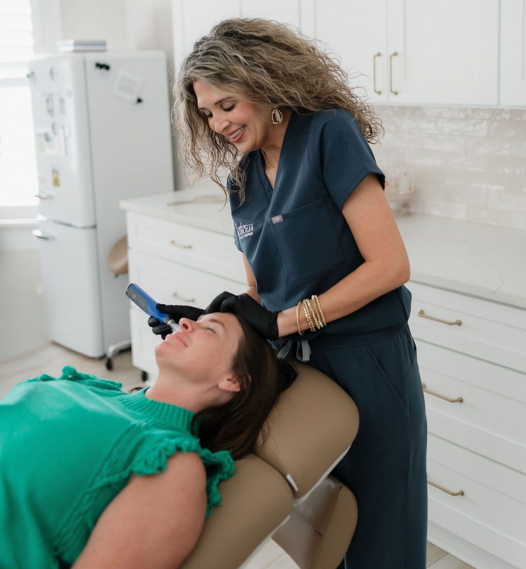 A woman clinician in navy scrubs performing a facial treatment with a handheld device on a reclining woman patient