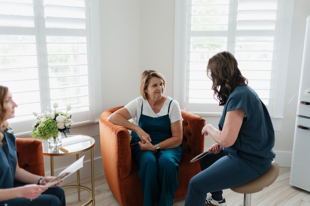 Two healthcare professionals consult with an older woman seated in an orange armchair in a bright clinic.