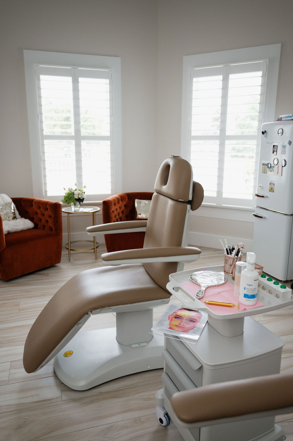 Bright aesthetic treatment room with beige reclining facial chair, trolley of skincare tools, and tufted orange lounge chairs.