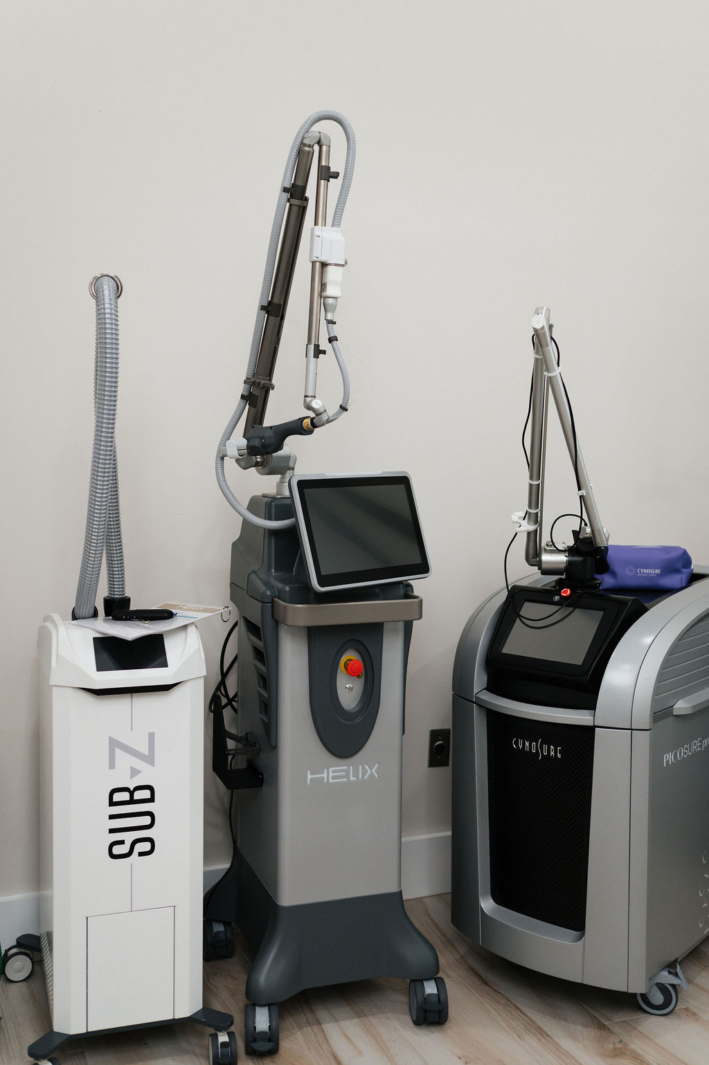 Three cosmetic laser machines (SUB-Z, HELIX, PicoSure) on rolling carts in a medical aesthetics treatment room