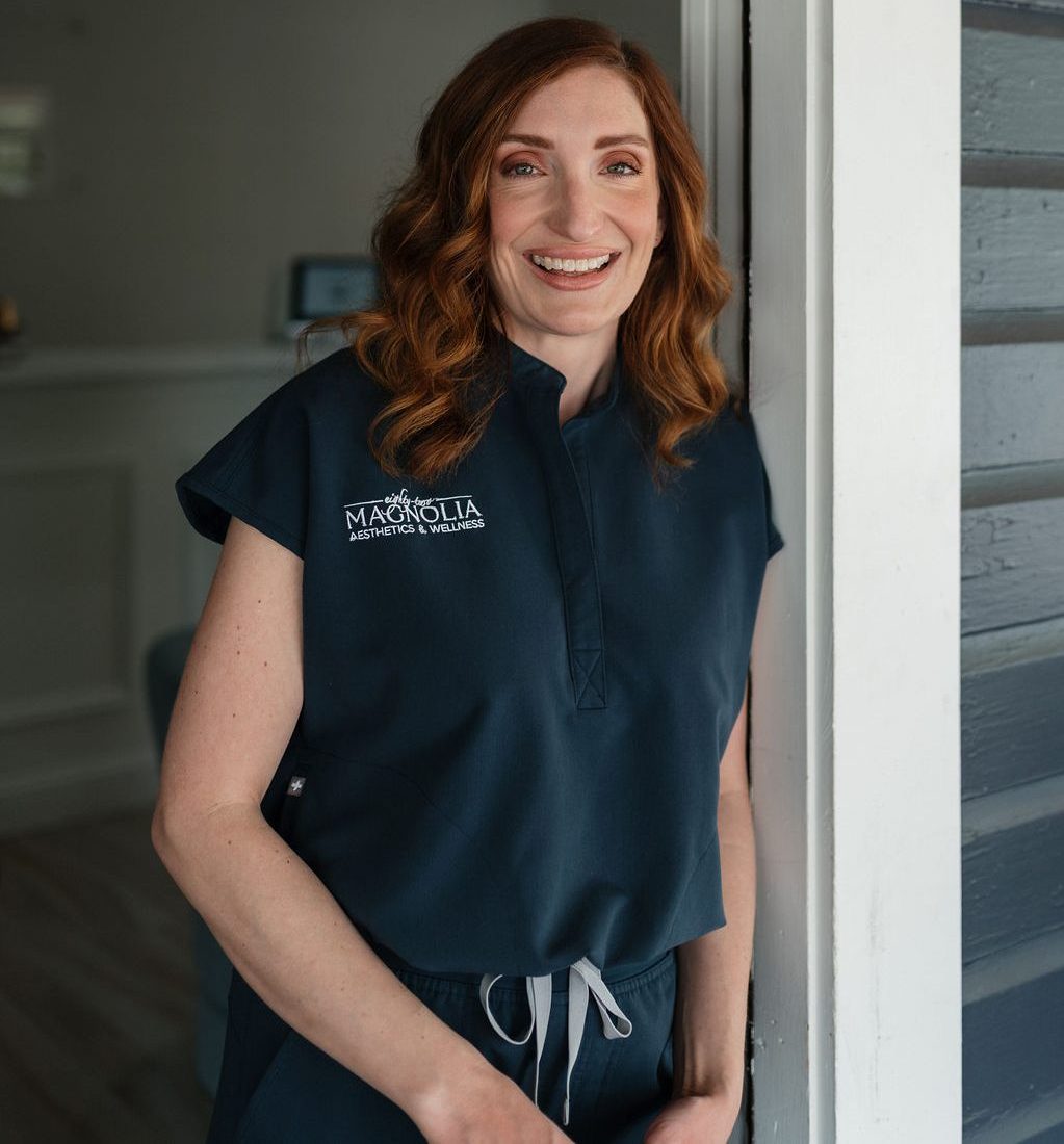 A smiling woman in navy scrubs embroidered Magnolia Aesthetics & Wellness leaning in a doorway.