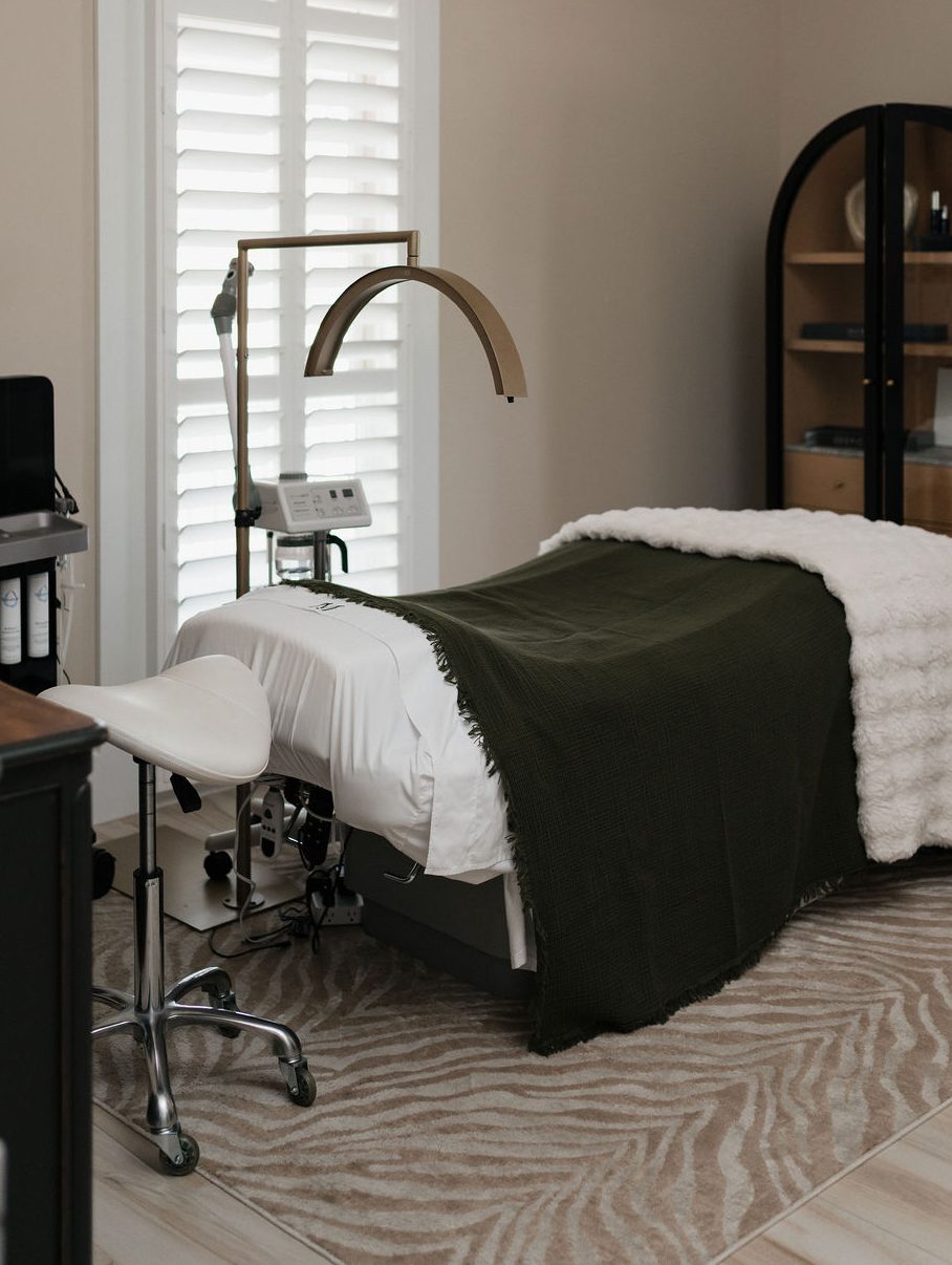 Calm spa treatment room with a massage table draped in dark green blanket, adjustable lamp, stool and shuttered window.