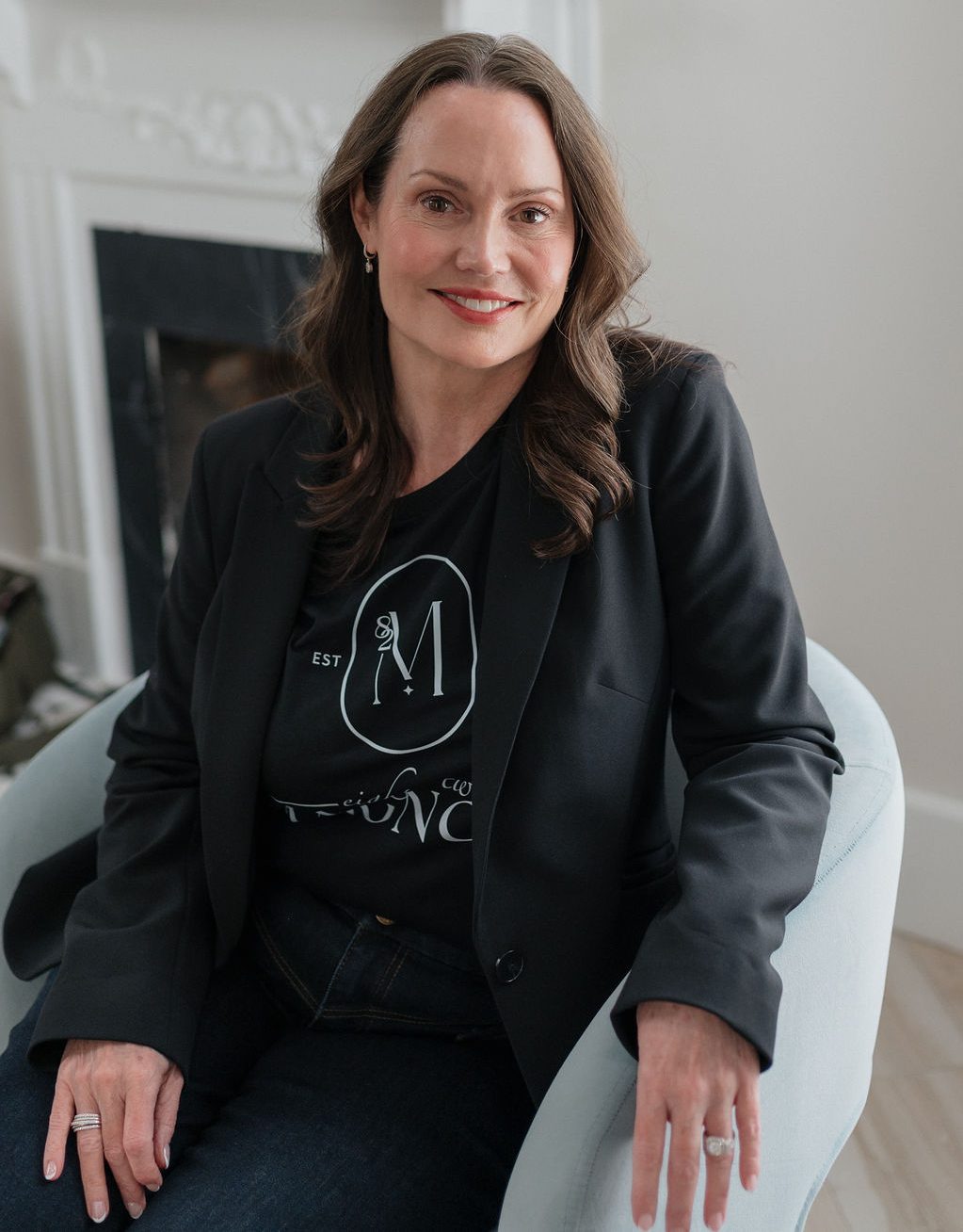 Smiling woman in a black blazer seated in a light living room armchair, fireplace and flowers in background.