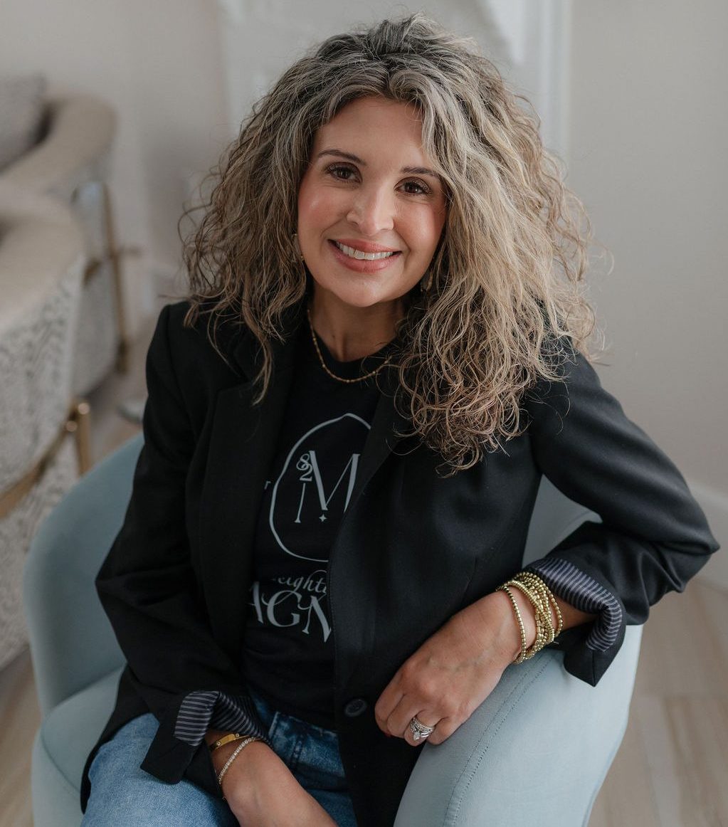 Smiling woman with curly gray hair wearing a black blazer and gold bracelets, seated in a bright living room.
