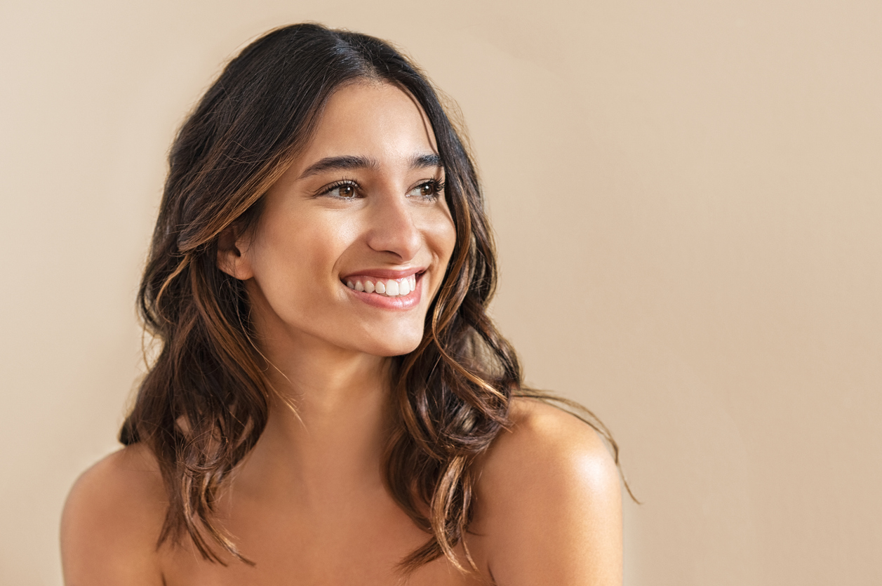 Smiling woman with wavy brown hair in natural light, close-up portrait on a beige background — skincare or beauty concept