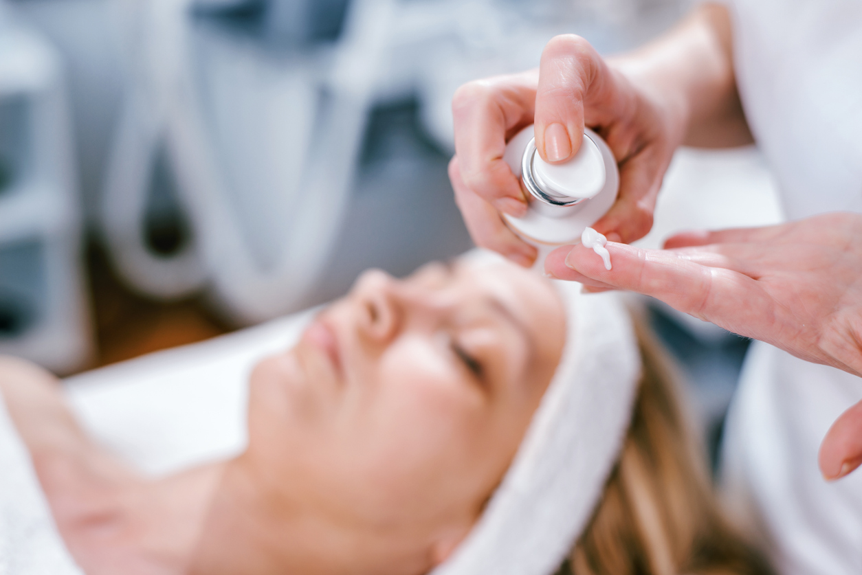 Esthetician dispensing facial cream onto finger before applying moisturizer to a relaxed woman at a spa facial.