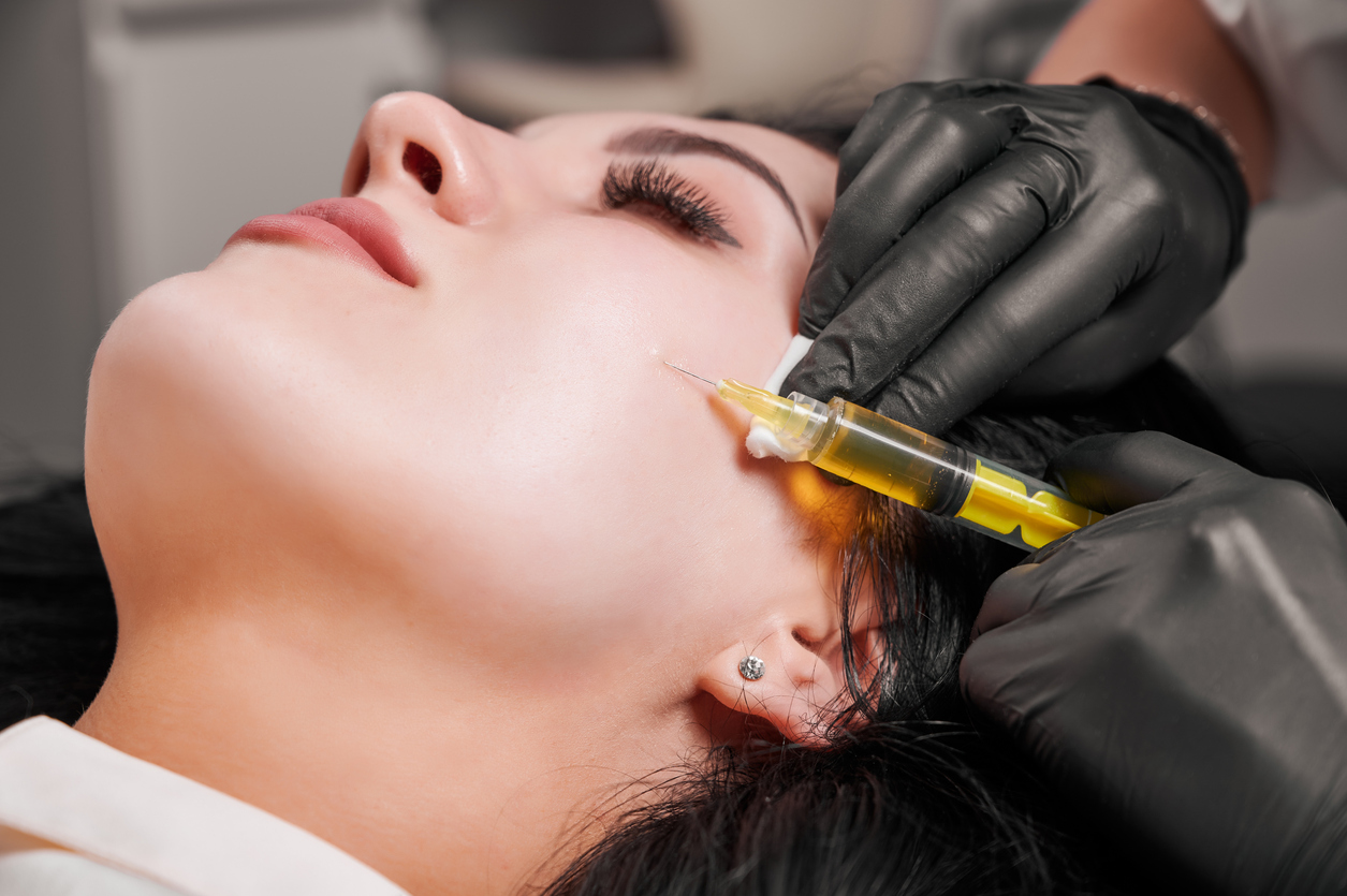 A woman receiving a cosmetic cheek injection from a practitioner wearing black gloves and a yellow-filled syringe.