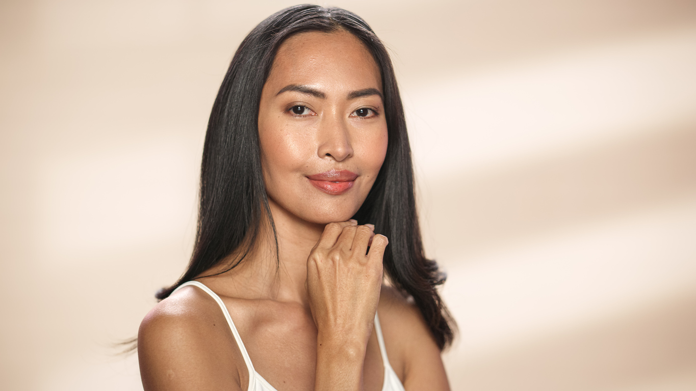 Portrait of a woman with long dark hair and natural makeup, hand on chin, wearing white tank top against beige background