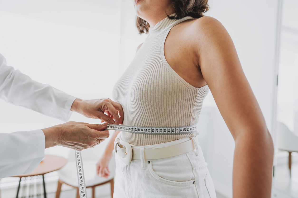 A woman in a ribbed tank top getting her bust measured with a tape measure by another person