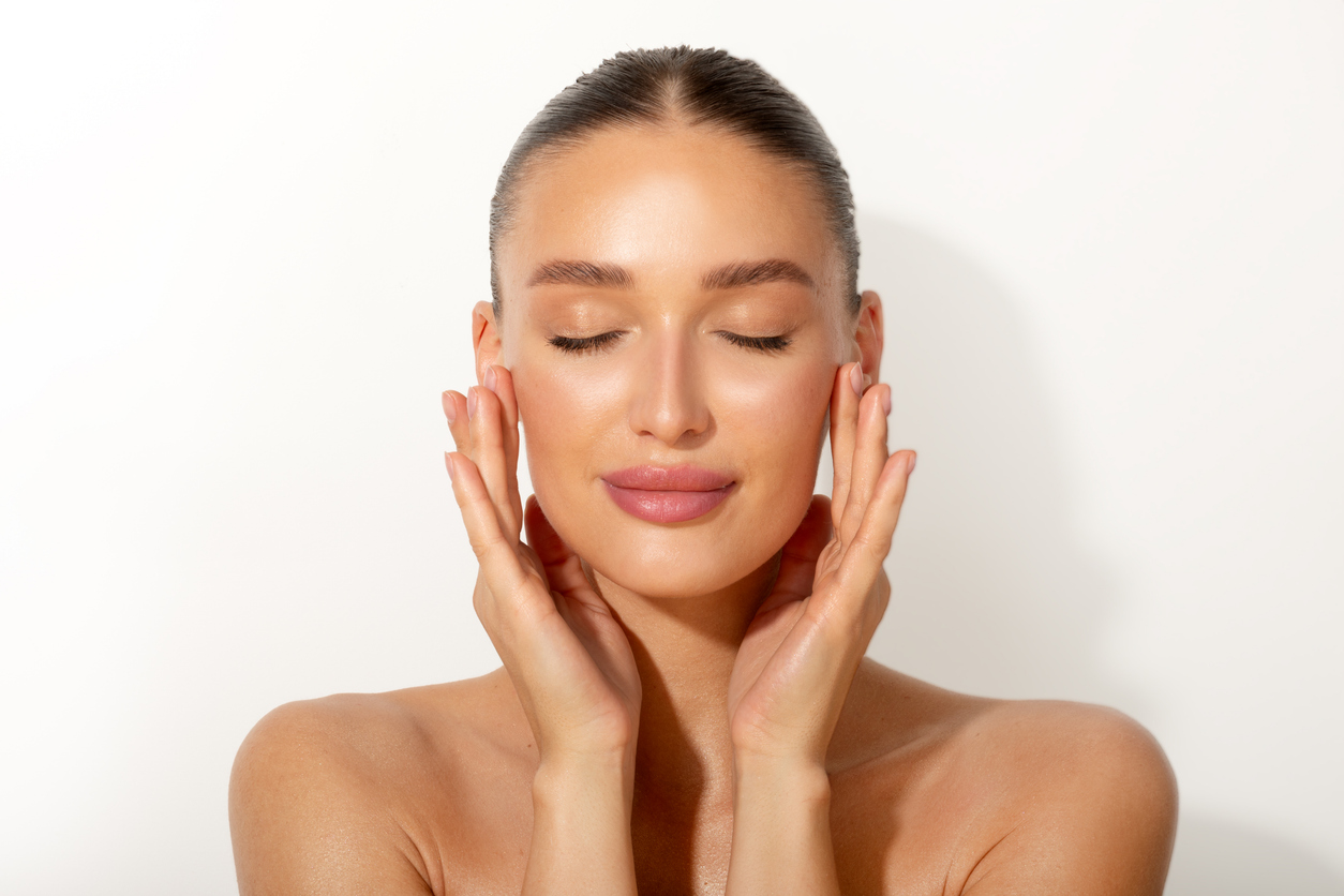 Young woman with glowing skin, eyes closed, hands gently touching face in a skincare beauty portrait