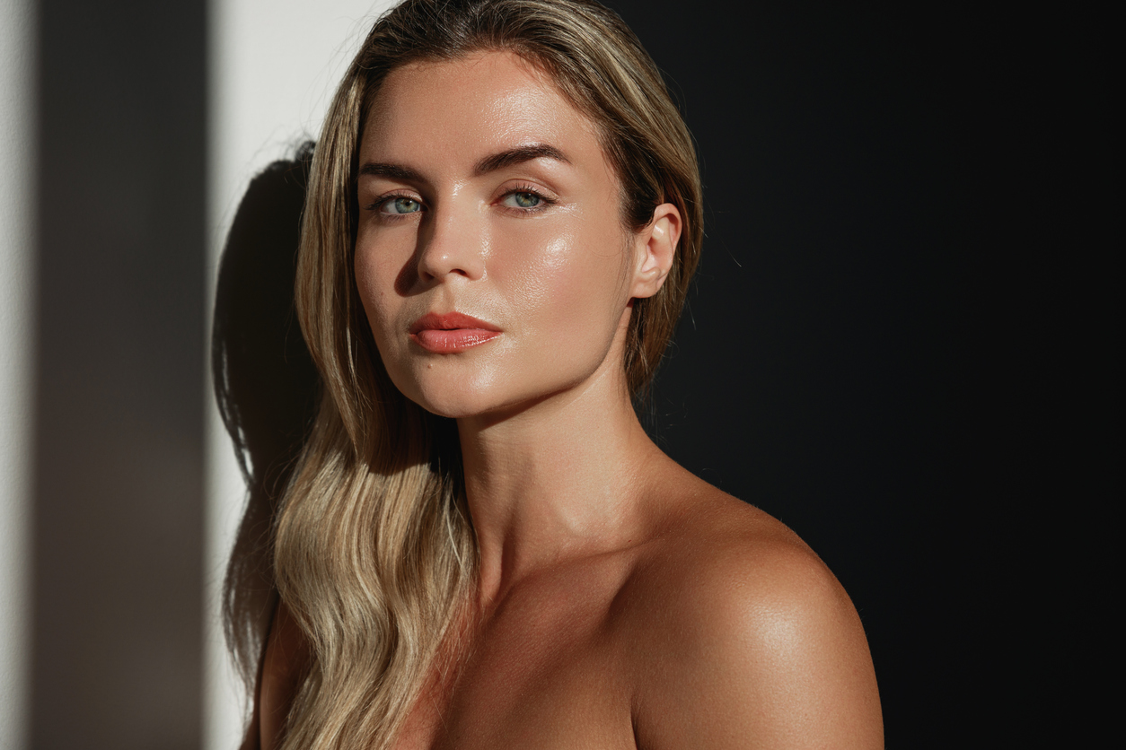 Sunlit portrait of a woman with blonde hair, glowing dewy skin and natural makeup against a dark background