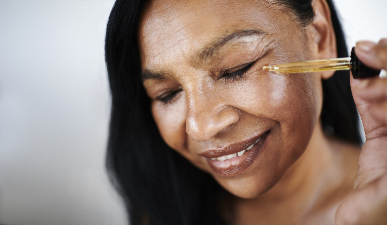 Middle-aged woman smiling while applying facial oil serum with dropper to cheek and eye area