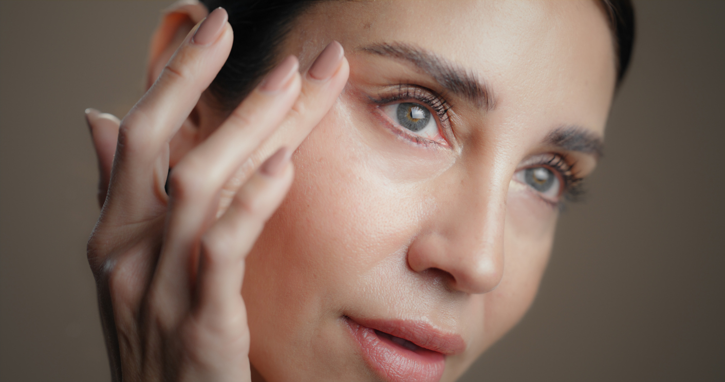 Close-up of a woman applying eye cream to under-eye, showing smooth skin, long lashes, and natural makeup.