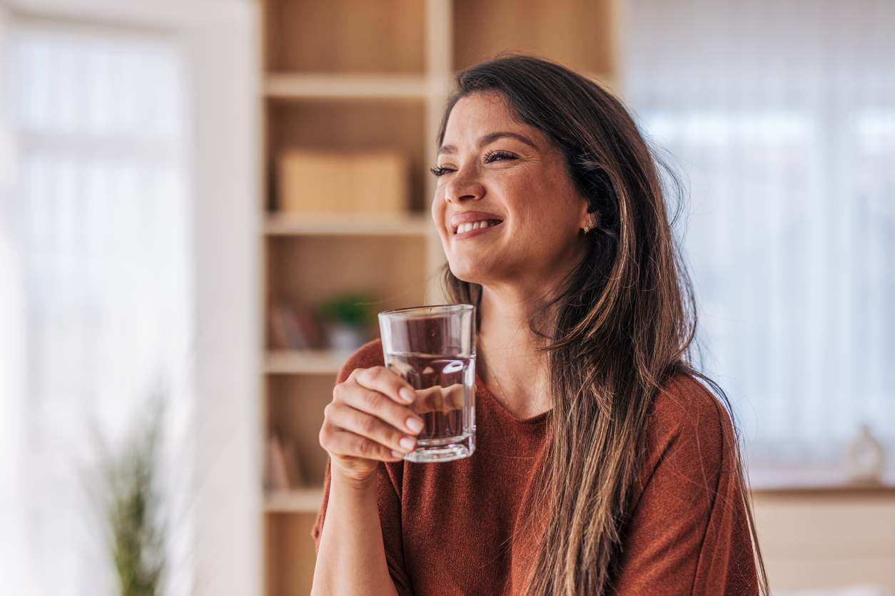 Smiling woman holding a glass of water in a cozy sunlit room, looking off-camera.