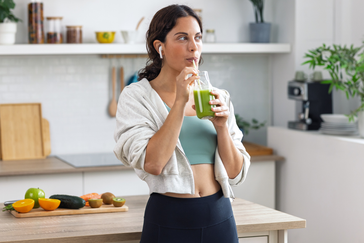 a woman in workout clothes sipping a green smoothie with earbuds in a bright kitchen, fruits on the counter