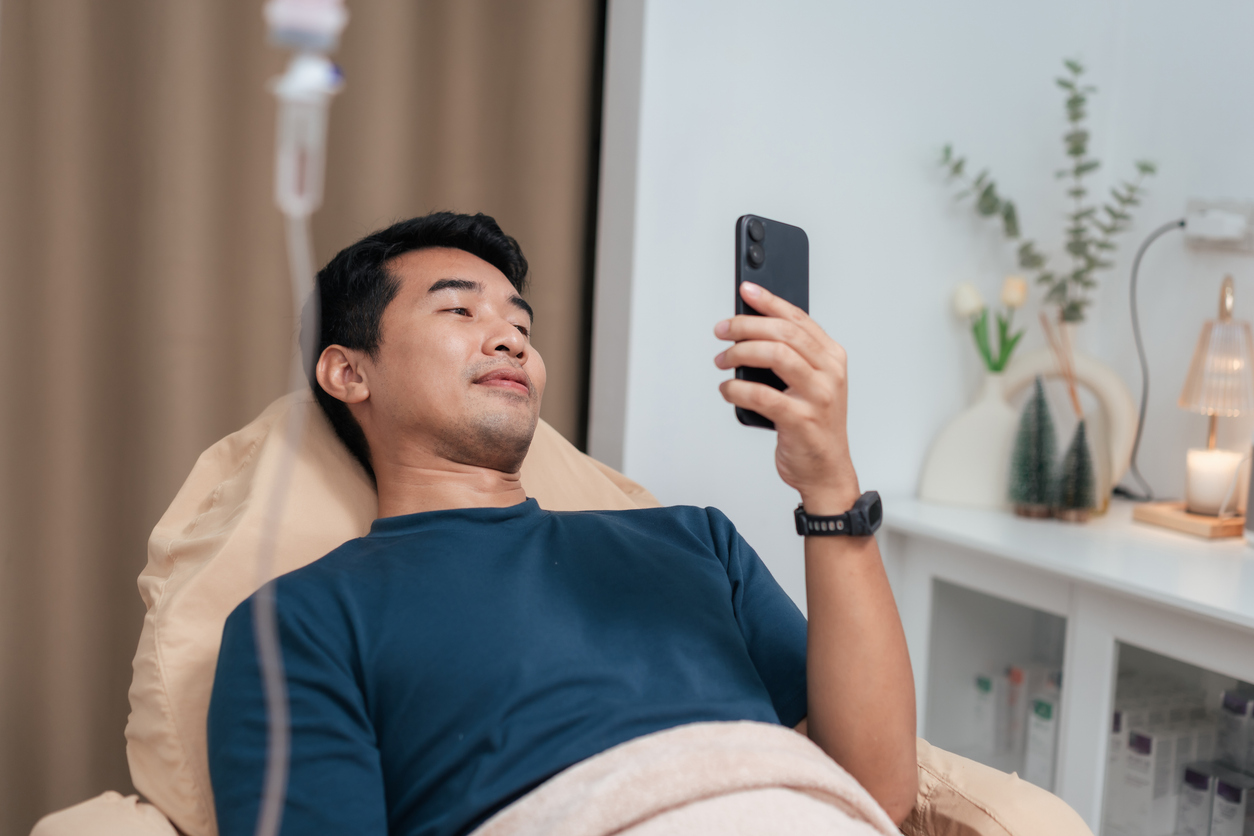 A man reclining in a clinic chair receiving IV therapy while using a smartphone