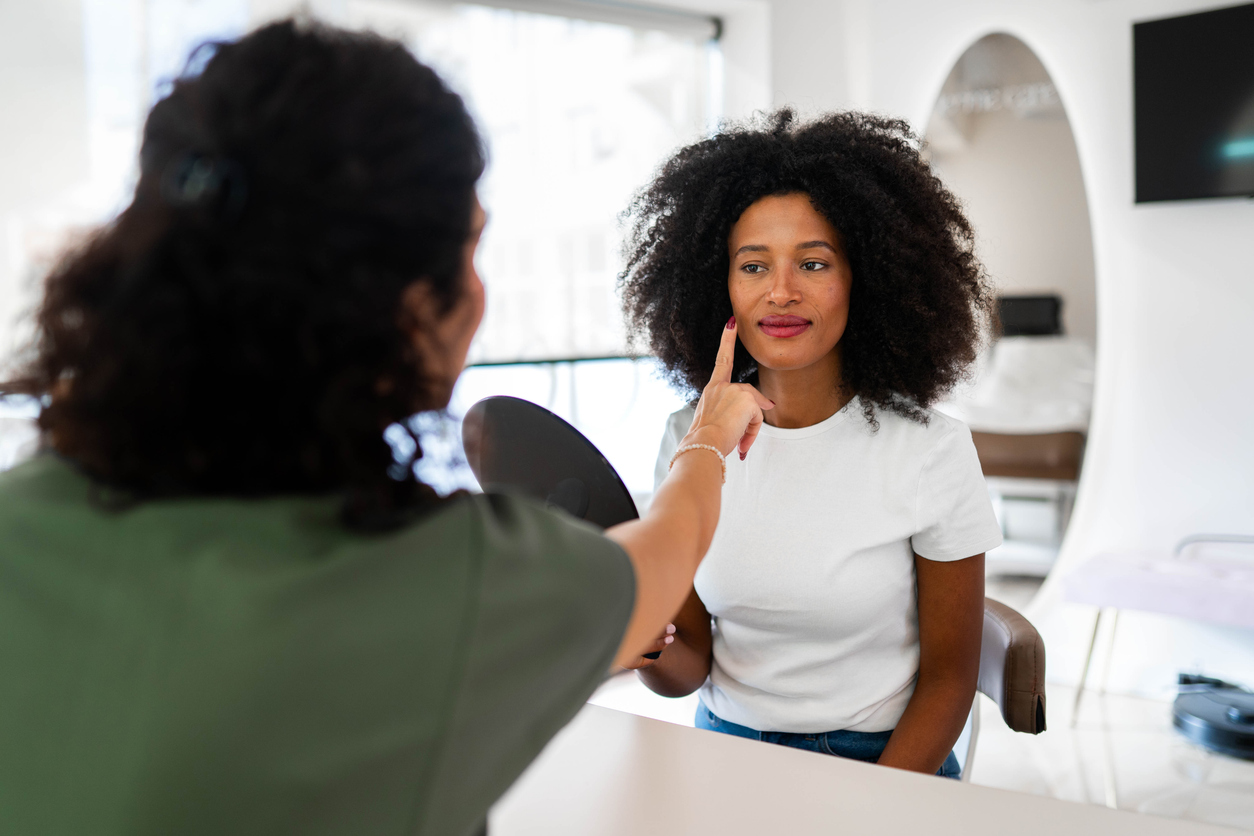 A woman at a skincare consultation, a specialist pointing to her cheek while she checks her face in a hand mirror