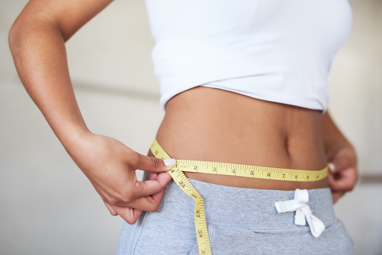 A woman measuring her waist with a yellow tape measure, wearing a white crop top and gray sweatpants